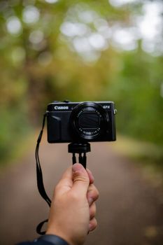 London, UK - September 30, 2020: Portrait View of a Hand Holding a Camera that faces the photographer, with a Blurry Forest as Background