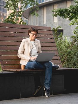 Freelance business woman sits in park with laptop and take away cardboard cup of coffee. Casual clothes, urban lifestyle of millennials. Working remotely.