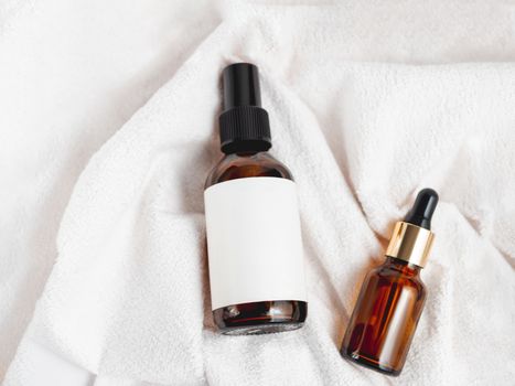 Top view on two glass bottles on white fluffy towel. Essential oil and cosmetic serum with clear labels on crumpled bathrobe