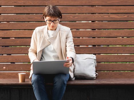 Business woman with short haircut and eyeglasses sits on wooden bench in park with laptop and cardboard cup of coffee. Student learns remotely from outdoors. Modern Internet technologies. Video call.