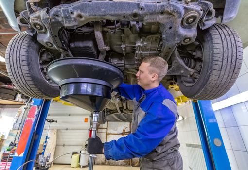 Happy car mechanic changes oil in a workshop. Mechanic standing under the car and draining oil into a special cannister via funnel.