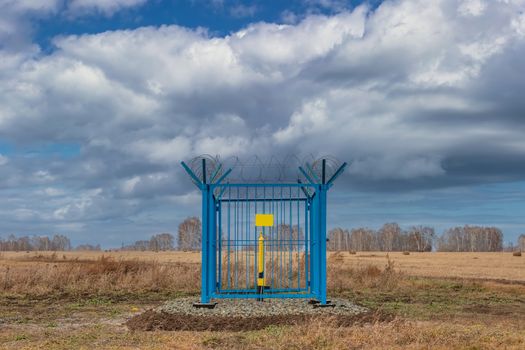 Shot of a caged industrial gas station with barbwire on top of the fence. Yellow gas pipe in the middle of the cage, empty yellow plate on the fence. Blue cloudy sky as a background. Siberia, Russia.