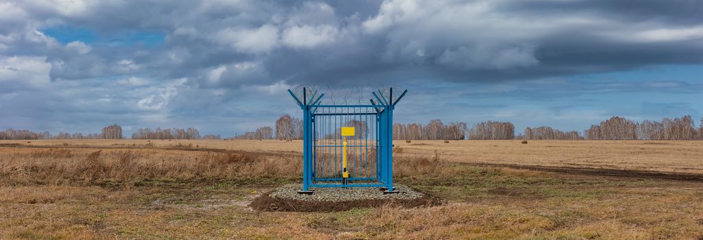 Panoramic shot of a caged industrial gas station with barbwire on top of the fence. Yellow gas pipe in the middle of the cage, empty plate on the fence. Cloudy sky as a background. Siberia, Russia.