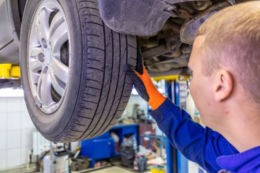 Close-up shot of mechanic checking very attentively surface of a car tire. Mechanic wearing blue coverall and orange gloves. Car is on a hydraulic lift.