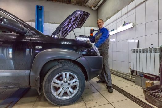 An auto mechanic standing next to a car with an open hood in a garage. He is holding a wrench on his shoulder, looking in the camera and smiling.