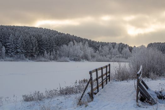 Wooden entrance to the forest near the frozen lake near the forest, all covered in snow. Dramatic sky with clouds over the winter forest and lake. Winter and frosty nature.