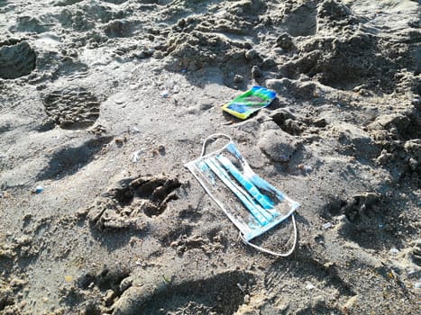 A blue mouthmask thrown on the sand of the beach with the coloured plastic sac of handkerchiefs in hard autumnal light and human footprint