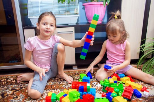 Children at home play with a plastic colored construction set. The girl built a tower from parts of a children's designer