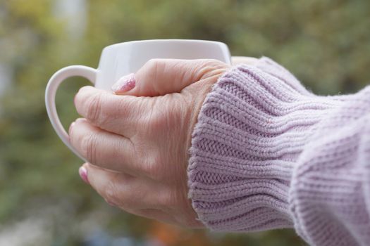 female hands in a warm sweater hold a hot cup on a nature background close-up