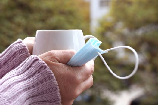 female hands hold a hot cup and medical mask on a nature background close up