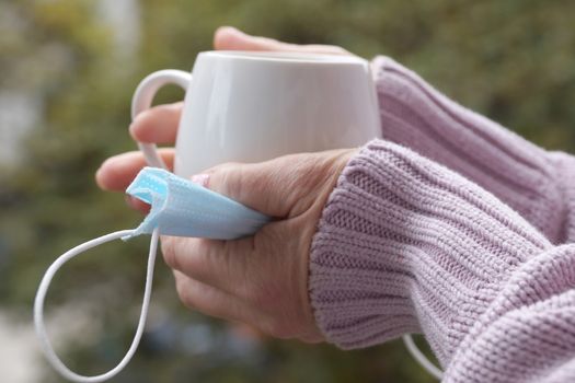 female hands hold a hot cup and medical mask on a nature background close up