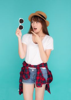 Beautiful portrait young asian woman wear hat and holding and pointing sunglasses with excited journey summer trip in vacation isolated on blue background, tourist asia girl surprise with travel.