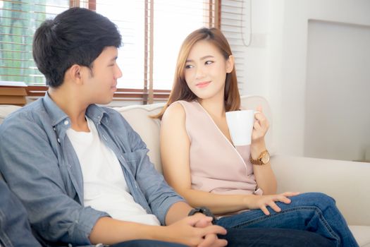 Beautiful young asian couple smiling and talking story married sitting on sofa at home together with woman holding a cup of coffee, man and woman relax and enjoy, lifestyle concept.