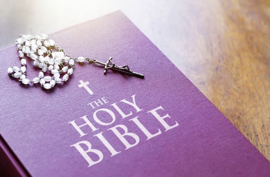 A rosary of white beads with a silver crucifix above the cover of the closed holy bible book. Religion and hope. Religious objects for praying