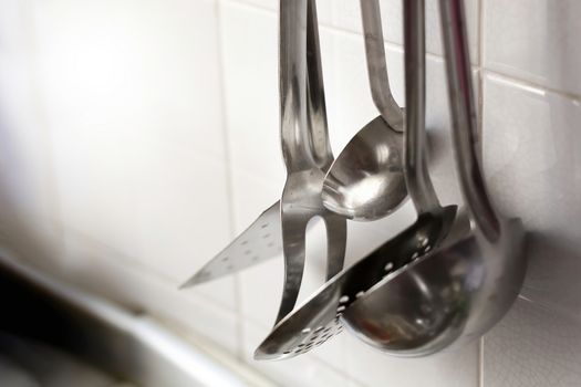 A skimmer, two forks and two metal ladles hanging on the tiled kitchen wall. Metal kitchen utensils