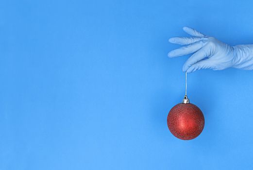 Hand in medical glove holds red christmas ball on a blue background with copy space.