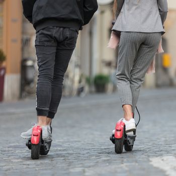 Rear view of unrecognizable trendy fashinable teenagers riding public rental electric scooters in urban city environment. New eco-friendly modern public city transport in Ljubljana, Slovenia.