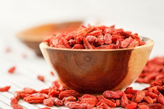 Dried goji berries in small wooden bowl, some scattered over white boards table under.