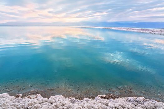 Salt crystals covering sand shore of Dead Sea, calm clear water surface near, typical morning scenery at Ein Bokek beach, Israel.
