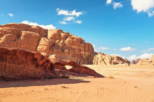 Rocky massifs on red sand desert, small stone arc bridge, bright blue sky in background - typical scenery in Wadi Rum, Jordan.