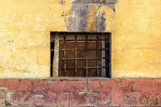 Old yellow wall with closed window with old rusty iron grid front