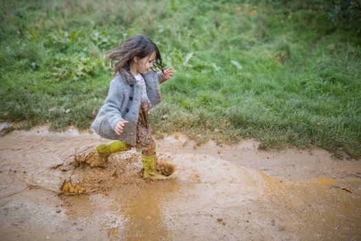 Happy little girl joyfully splashing and running through a muddy puddle. Young child having fun and getting dirty in puddles. Kids playing outside