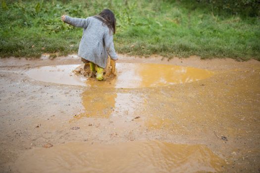 Happy little girl on dirt road joyfully jumping and splashing in muddy puddle. Young child having fun jumping in puddles. Kids playing outside