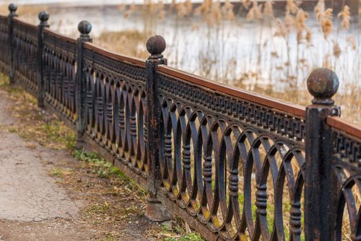 old rusted falling apart cast iron russian fence with ball ends and cracked asphalt in front of abandoned lake with selective focus blur