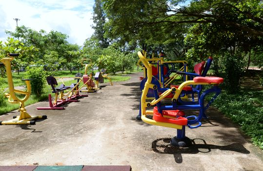 The colorful outdoor exercise machines in the park are lush many green trees.