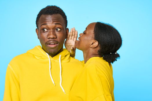 A woman whispers to a man in the ear of a young couple of African appearance in yellow sweaters on a blue background