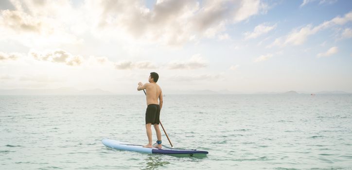 Asian man is paddling on a SUP board, standup paddler at the ocean during sunset, summer holidays vacation travel.