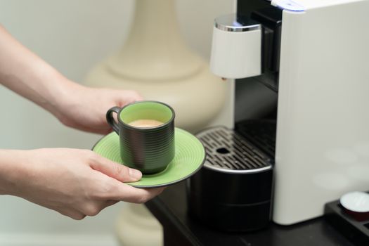 Machine serving coffee in a cup. Coffee capsule machine maker. Hand holding a ceramic cup of coffee on the coffee machine.