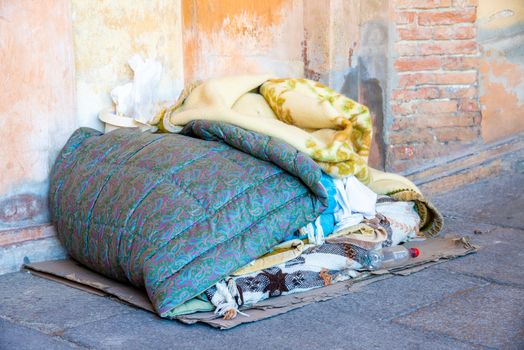 The sleeping place of a homeless man with blankets cleared together, belongings on the ground in front of a wall with chipped plaster in a city