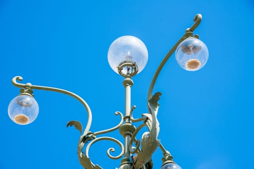 A street lamp in Verona, located in the historic center, artistically adorned with wrought-iron figures and forms and yet operated with halogen light, photographed against a blue sky.