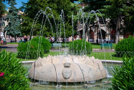 The fountain "Fontana di Piazza Brà" in Verona Italy on a busy square with many plants around and bubbling water in the sun.