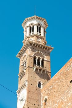A church tower in Verona photographed from the outside and diagonally below made of stone, a clock, with several ledges in front of a blue sky
