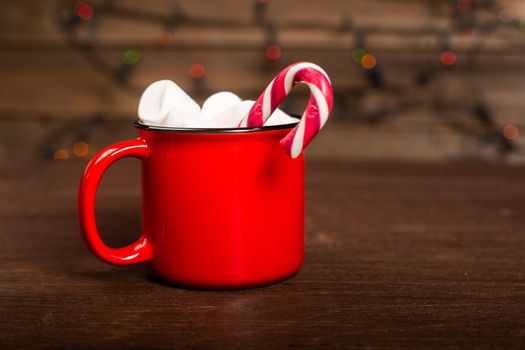 Cocoa in red mug with marshmallows and candy cane on dark wooden background with bokeh lights