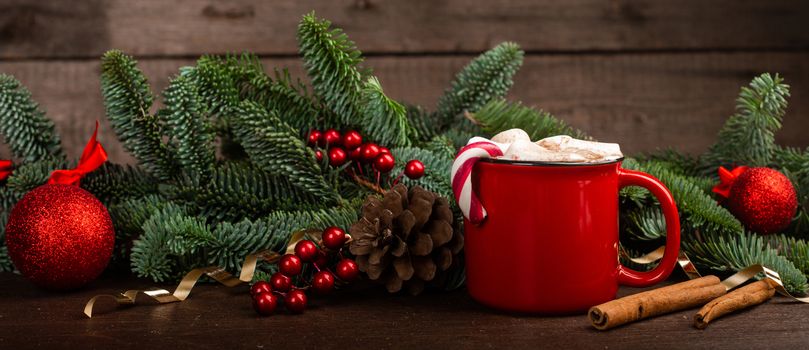 Cocoa hot chocolate in red mug with marshmallows candy cane fir tree branches and red baubles on dark wooden background