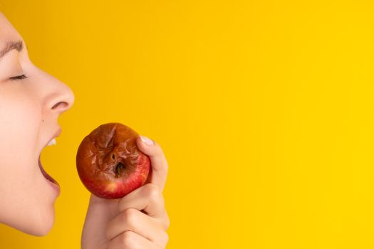 The girl bites a rotten apple with a worm on a yellow background. Expired products, junk food.