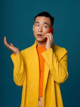 A fashionable man in a yellow jacket and an orange T-shirt is talking on the phone on a blue background. High quality photo
