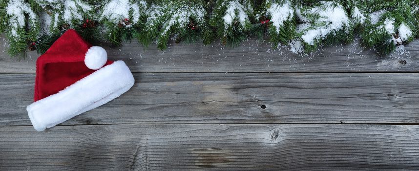 Merry Christmas concept on rustic natural wooden background with snow covered Fir branches plus Santa Claus cap   