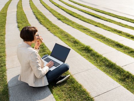 Freelance business woman sits in park with laptop and take away cardboard cup of coffee. Casual clothes, urban lifestyle of millennials. Working remotely.
