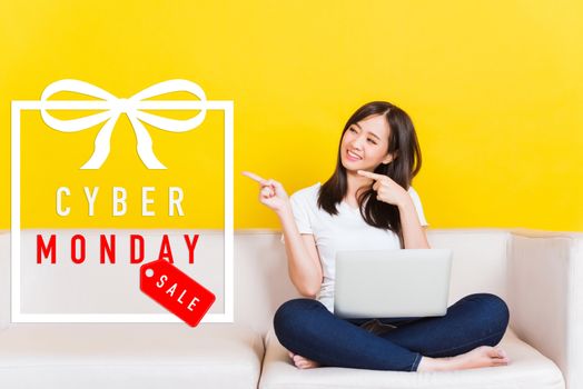 Asian happy beautiful young woman she sitting on sofa using laptop computer shopping online in house living room point finger to Cyber Monday text in gift box isolated on yellow background
