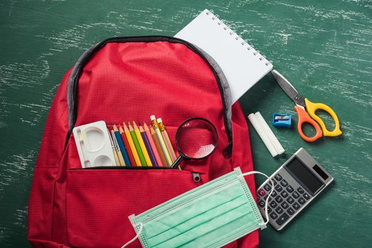 Top view of stylish red school bag backpack at a green chalkboard with face mask protection and stationery, Back to school education new normal during outbreak COVID-19 or coronavirus concept