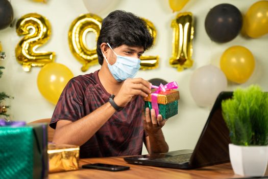 Young man with medical mask opening gift box over video call on laptop with 2021 new year decorated background - concept of distant xmas or holiday celebration due to covid-19 or coronavirus pandemic