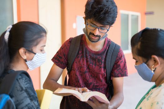 Group of college students in medical mask discussing by looking into book at college campus corridor - concept of college reopen, togetherness and friendship