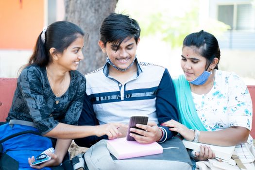 Three friends while sitting at college laughing by looking into mobile phone - concept of students internet, mobile addiction, online bullying or gossips at university campus.