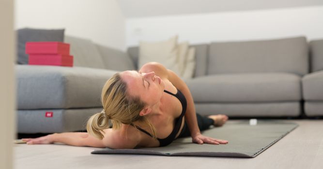 Beautiful blonde woman doing home workout indoors. Woman practice yoga at home. Fit girl using workout tutorials for healthy active lifestyle. Woman using quarantine for home workouts. Stretching