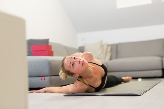 Beautiful blonde woman doing home workout indoors. Woman practice yoga at home. Fit girl using workout tutorials for healthy active lifestyle. Woman using quarantine for home workouts. Stretching