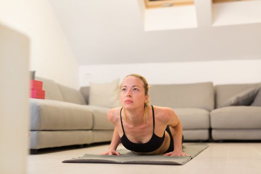 Beautiful blonde woman doing home workout indoors. Woman practice yoga at home. Fit girl using workout tutorials for healthy active lifestyle. Woman using quarantine for home workouts. Stretching